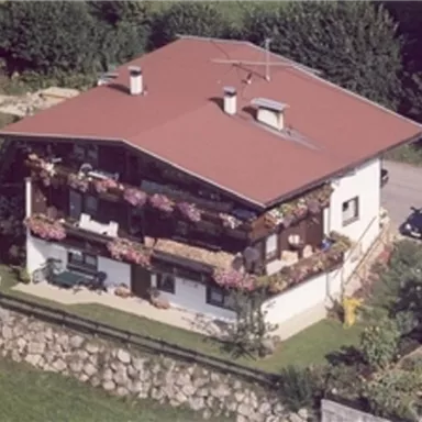 Ein hübsches, zweigeschossiges Haus mit einem roten Dach und einem Balkon voller Blumen. Es liegt in einer grünen Umgebung, umgeben von Bäumen.