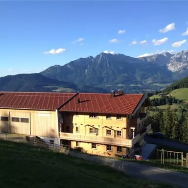A picturesque mountain landscape with a wooden building in the foreground. The surroundings are green and the mountains are majestically visible in the background.