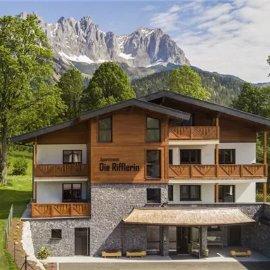 A modern building with a wooden facade, surrounded by green grass and trees. In the background, majestic mountains can be seen.