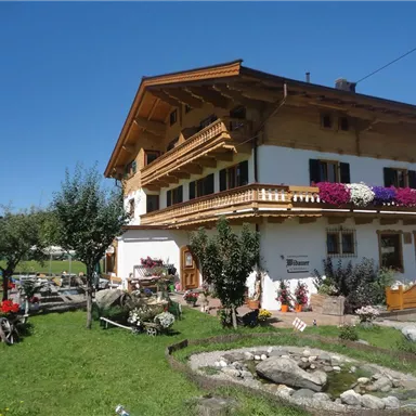 Ein charmantes Holzhaus mit traditioneller Architektur und bunten Blumen an den Fenstern. Der Garten ist gepflegt und es gibt einen kleinen Teich mit Steinen.