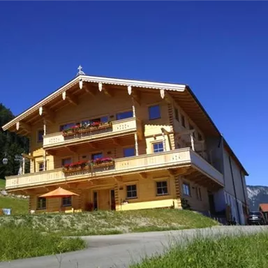 A charming wooden house with balconies and colorful flowers. Surrounding it are green meadows and mountains in the background.