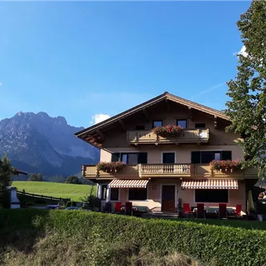 Ein charmantes Berghaus mit Balkon und Blumen. Im Hintergrund erheben sich majestätische Berge unter einem klaren blauen Himmel.