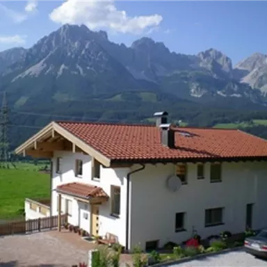 A beautiful house with a red roof in a green landscape. In the background, majestic mountains rise under a blue sky.
