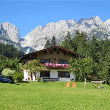 A pretty house in the mountains, surrounded by trees and meadows. In the background, majestic mountain peaks rise under a clear blue sky.