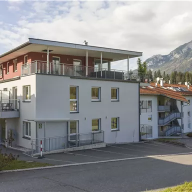 A modern building with several apartments and a flat roof. In the background, mountains and a clear sky can be seen.