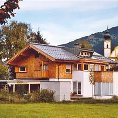 Ein modernes Haus mit Holzfassade und großem Fenster. Im Hintergrund sind Berge und eine Kirche sichtbar.