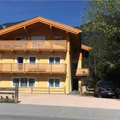 Ein gelbes Gebäude mit Holzverkleidung und mehreren Balkonen. Es befindet sich in einer ruhigen Umgebung mit Bäumen und Bergen im Hintergrund.