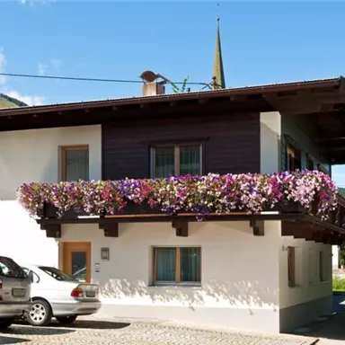 A charming house in alpine style with a wooden balcony and colorful flowers. In the background, gentle hills and blue sky can be seen.