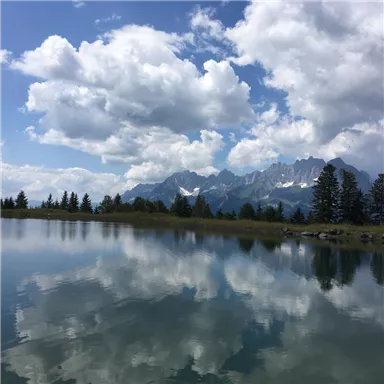 Eine ruhige Landschaft mit einem klaren See und majestätischen Bergen im Hintergrund. Die Wolken spiegeln sich sanft im Wasser.