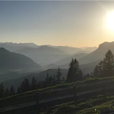 Eine malerische Berglandschaft mit sanften Hügeln und einem strahlenden Sonnenuntergang. Im Vordergrund sind Bäume und ein Holzgeländer sichtbar.