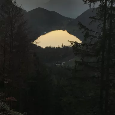 Eine malerische Berglandschaft bei Sonnenuntergang. Ein ruhiger See spiegelt die Farben des Himmels wider, umgeben von Wäldern.