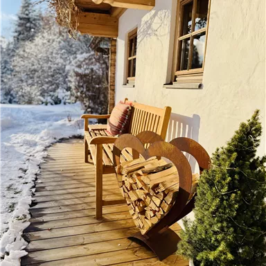 A cozy wooden bench with a heart motif, storing wood for the fireplace. The path is surrounded by fresh snow, and in the background, there are snow-covered trees.