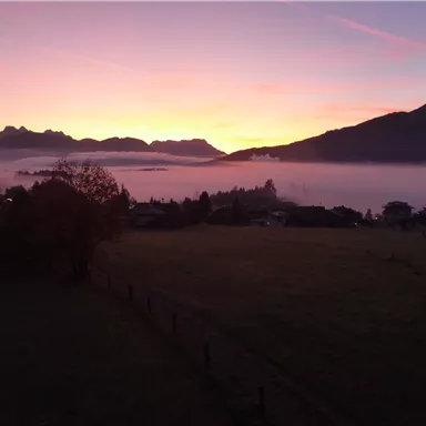 A picturesque landscape at sunrise with mountains in the background. The sky is painted in soft colors, while fog drifts over the valley.