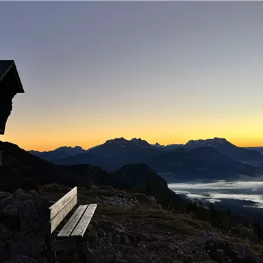 Eine Bank steht auf einem Berghang mit Blick auf die Berge bei Sonnenaufgang. Der Himmel ist in warmen Farben und Nebel zieht über das Tal.