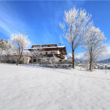 Ein Winterhaus umgeben von schneebedecktem Gelände und gefrorenen Bäumen. Der Himmel ist klar und strahlt in Blau.