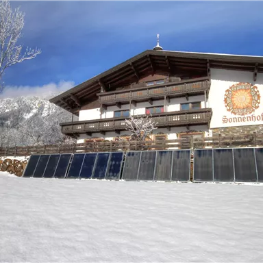 Ein Fachwerkhaus im Schnee mit Solarpanels im Vordergrund. Im Hintergrund sind Berge und ein blauer Himmel zu sehen.