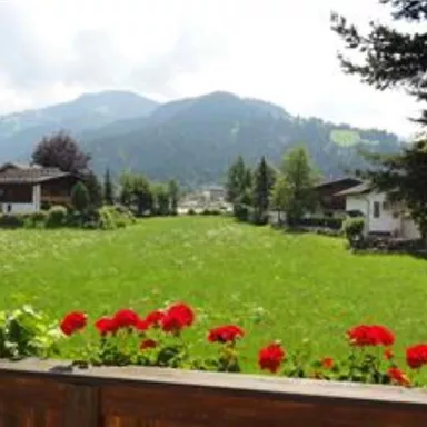A picturesque landscape with green meadows and mountains in the background. Red flowers adorn the foreground and rustic houses are scattered around the meadow.