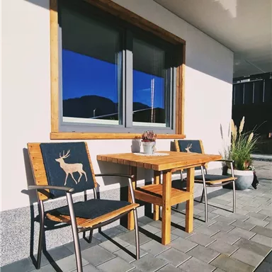 Ein gemütlicher Außenbereich mit einem Holztisch und zwei Stühlen. Die Stühle sind mit einem Reh-Motiv verziert und es gibt eine Topfpflanze im Hintergrund.