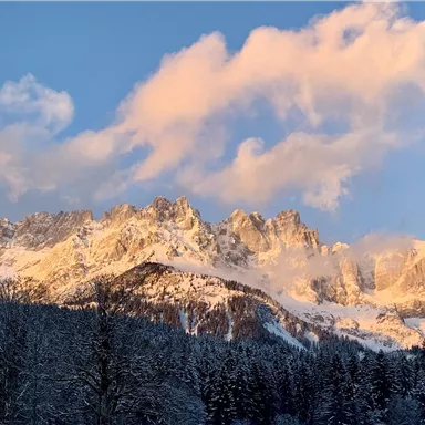 An impressive mountain landscape with snow-covered peaks and gentle clouds. The sun illuminates the mountains in warm colors.