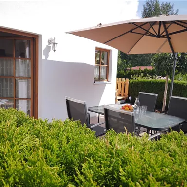 An inviting outdoor area with a table and chairs under a sunshade. Green hedges surround the space and provide privacy.