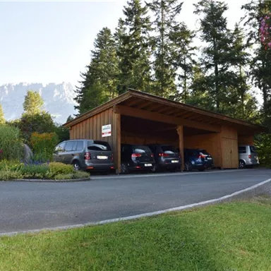 Eine überdachte Parkfläche mit mehreren Autos steht umgeben von grünen Wiesen und Bäumen. Im Hintergrund sind Berge zu sehen.