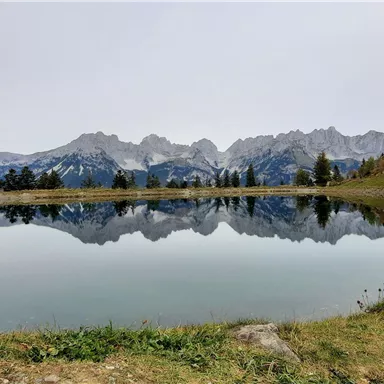A calm lake reflects the surrounding mountains. The landscape is surrounded by gentle green meadows.