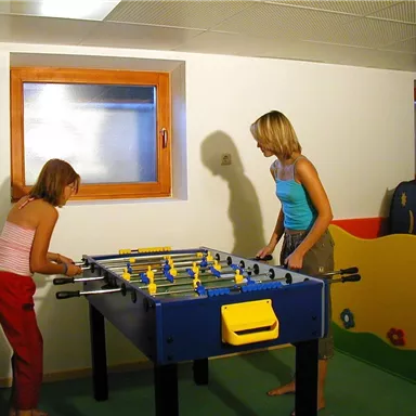 Zwei Kinder spielen Tischfußball in einem hellen Raum. Im Hintergrund sind bunte Möbel zu sehen.
