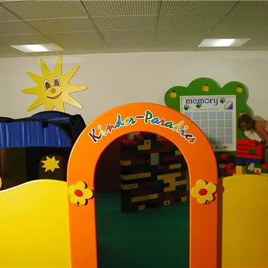 Ein farbenfroher Spielbereich mit einem Bogen, der "Kinder-Paradies" zeigt. Im Hintergrund sind eine Sonne und verschiedene Spielstationen zu sehen.