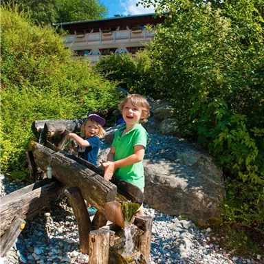 Zwei fröhliche Kinder spielen an einer Wassermühle in der Natur. Im Hintergrund sind Bäume und ein Gebäude zu sehen.