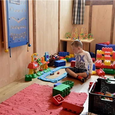 Ein kleiner Junge spielt in einem bunten Spielzimmer mit verschiedenen Spielzeugen und Bauklötzen. Der Raum ist gemütlich mit Holzwänden und einem roten Teppich.