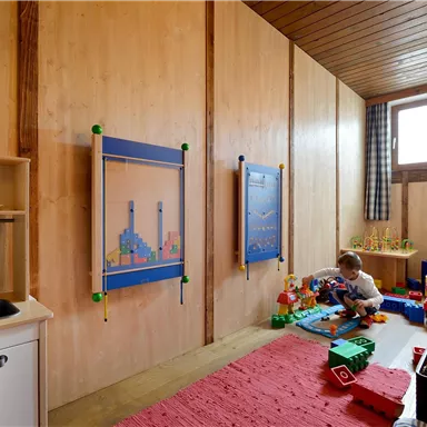 Ein heller Spielraum mit Holzverkleidung und bunten Spielzeugen. Ein Kind spielt mit Bausteinen auf einem roten Teppich.