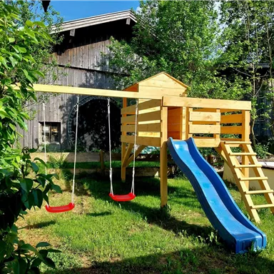 A playground with a slide and two swings is located in the green garden. In the background, you can see a wooden structure and a lot of fresh foliage.