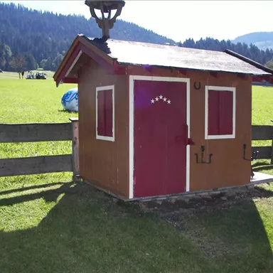 Ein kleines, bunten Häuschen steht auf einer grünen Wiese. Im Hintergrund sind Berge und eine Holzbank zu sehen.