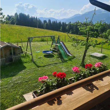 Ein schöner Garten mit einer Schaukel, Rutsche und einer Rasenfläche. Im Hintergrund sind grüne Wiesen und Berge zu sehen.