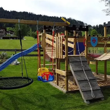 Ein Spielplatz aus Holz mit einer Rutsche, Schaukeln und einem Klettergerüst. Die Umgebung ist grün mit Bergen im Hintergrund.