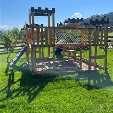 Ein Holzspielplatz mit einer Rutsche und einer Netzschaukel. Er steht auf grünem Gras unter blauem Himmel.
