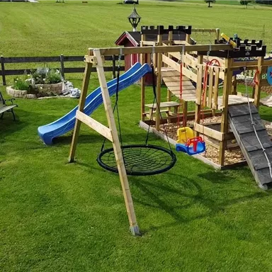 Ein schöner Spielplatz mit einer Holzspielburg, einer Rutsche und einer Schaukel. Daneben steht ein Trampolin auf dem grünen Rasen.