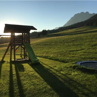 A playground with a slide and swings is located on a green meadow. In the background, there are mountains and a clear sky.