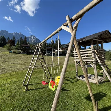 Ein Spielplatz mit einer Holzschaukel und einer Rutsche auf einer grünen Wiese. Im Hintergrund sind Berge und ein blauer Himmel mit einigen Wolken zu sehen.