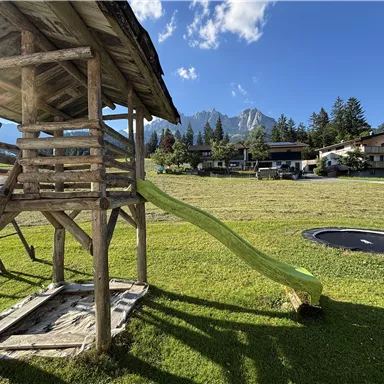 Ein Spielplatz mit einer Holzspielstruktur und einer Rutsche. Im Hintergrund sind grüne Wiesen und hohe Berge zu sehen.