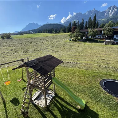 Ein Spielplatz mit einer Rutsche, Schaukeln und einem Trampolin umgeben von einer grünen Wiese. Im Hintergrund sind Berge und einige Gebäude zu sehen.