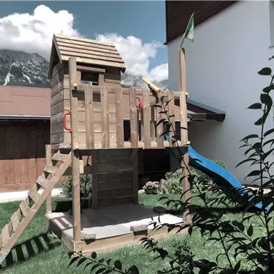 Ein Holzspielplatz mit einer Rutsche und einer Aussichtsplattform steht im Garten. Im Hintergrund sind Berge und eine helle Fassade eines Hauses zu sehen.