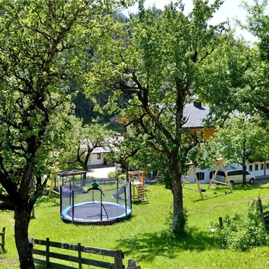 Ein schöner Garten mit vielen Bäumen und einer großen Trampolin. Im Hintergrund sind ein Spielplatz und ein Gebäude zu sehen.