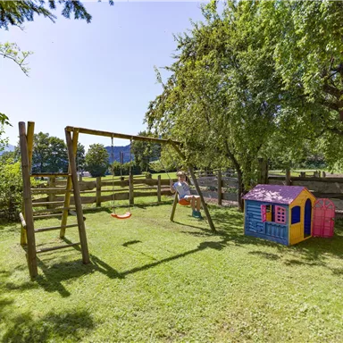 Ein schöner Spielplatz mit einer Schaukel und einem bunten Spielhaus. Die Umgebung ist grün und von Bäumen umgeben.