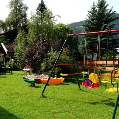 Ein schöner Garten mit einer Schaukel und einem Spielzeugwagen. Im Hintergrund sind Bäume und ein Haus zu sehen, umgeben von Bergen.