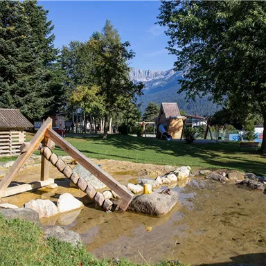 Ein Spielplatz mit einem Wasserbecken und einer kleinen Brücke aus Holz. Im Hintergrund sind Bäume und die Berge sichtbar.