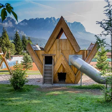 Ein moderner Spielplatz mit einem hölzernen Spielhaus in Form einer Spitze. Im Hintergrund sind Berge und Bäume sichtbar.