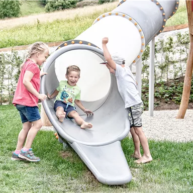 Three children are playing on a slide outdoors. One child joyfully slides down while the others watch and laugh.