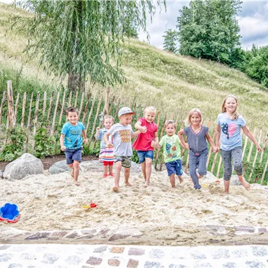 A group of seven children is playing in the sand. In the background, green meadows and trees can be seen.