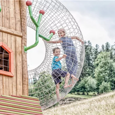 A playground with a wooden house and a large net ball. Two children are playing and having fun outdoors.